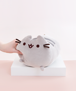 Model’s hand strongly squeezing the Medium Log Squisheen plush’s face, causing the left nub foot to move upwards. The plush has been placed on a square white pedestal in front of a light pink and white background. 