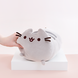 Model’s hand strongly squeezing the Medium Log Squisheen plush’s face, causing the left nub foot to move upwards. The plush has been placed on a square white pedestal in front of a light pink and white background. 