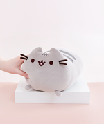 Model’s hand lightly squeezing the Medium Log Squisheen plush, which has been placed on a square white pedestal in front of a light pink and white background. The Medium Log plush features Pusheen laying on her stomach, shaped like a cylinder with two nub paws upfront. Pusheen’s eyes and slightly open mouth are embroidered onto her face, and she has two felt whiskers poking out from each side of her face. 