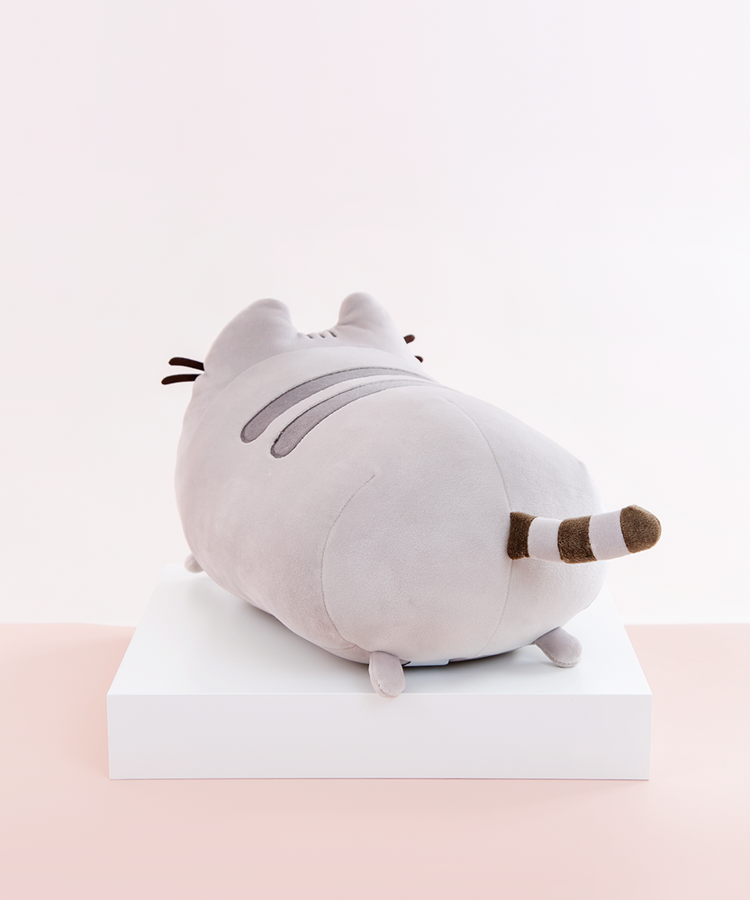 Back quarter view of the Medium Log Squisheen, facing the left, placed on top a square white pedestal in front of a light pink and white background. Pusheen’s behind is just as round as her front. Her striped tail sits directly in the middle of the circle. There are two nub feet at the bottom of the circle, mirroring the ones up front. 