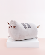 Side view of the Medium Log Squisheen, facing the left, placed on top a square white pedestal in front of a light pink and white background. Pusheen’s back feet and striped tail stick out from her behind.