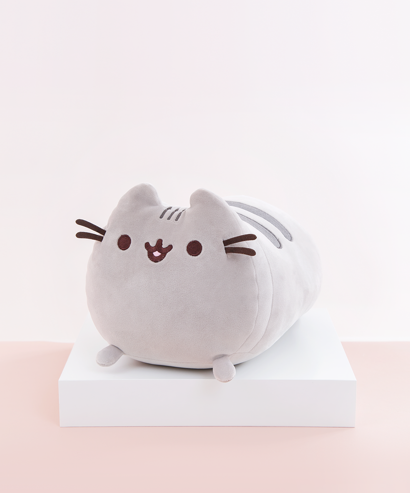 Quarter profile view of the Medium Log Squisheen, facing the left, placed on top a square white pedestal in front of a light pink and white background. The front of Pusheen is a perfect circle, save for her two ears poking out on top. Pusheen’s face and whiskers are located closer to the top, while her nub feet are at the very bottom. Pusheen’s body is shaped like a log, with two dark grey stripes on top of her back.