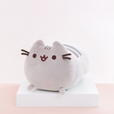 Quarter profile view of the Medium Log Squisheen, facing the left, placed on top a square white pedestal in front of a light pink and white background. The front of Pusheen is a perfect circle, save for her two ears poking out on top. Pusheen’s face and whiskers are located closer to the top, while her nub feet are at the very bottom. Pusheen’s body is shaped like a log, with two dark grey stripes on top of her back.