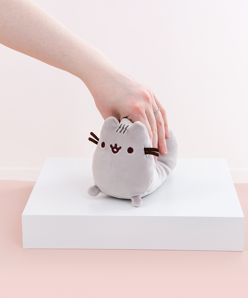 Model’s hand strongly squeezing the Mini Log Squisheen plush, which has been placed on a square white pedestal in front of a light pink and white background.