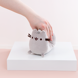 Model’s hand strongly squeezing the Mini Log Squisheen plush, which has been placed on a square white pedestal in front of a light pink and white background.