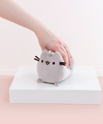 Model’s hand lightly squeezing the Mini Log Squisheen plush, which has been placed on a square white pedestal in front of a light pink and white background.