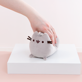 Model’s hand lightly squeezing the Mini Log Squisheen plush, which has been placed on a square white pedestal in front of a light pink and white background.