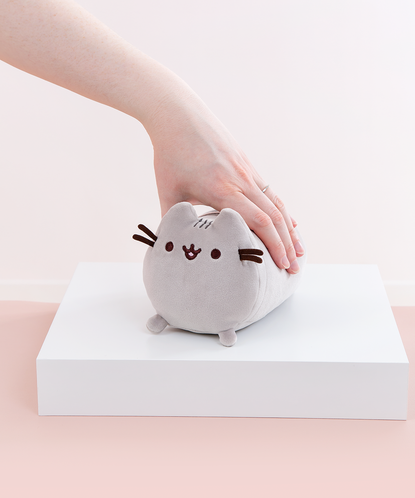 Model’s hand resting on top of the Mini Log Squisheen plush, which has been placed on a square white pedestal in front of a light pink and white background. The mini plush features Pusheen laying on her stomach, shaped like a cylinder with two nub paws upfront. Pusheen’s eyes and slightly open mouth are embroidered onto her face, and she has two felt whiskers poking out from each side of her face. 