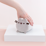 Model’s hand resting on top of the Mini Log Squisheen plush, which has been placed on a square white pedestal in front of a light pink and white background. The mini plush features Pusheen laying on her stomach, shaped like a cylinder with two nub paws upfront. Pusheen’s eyes and slightly open mouth are embroidered onto her face, and she has two felt whiskers poking out from each side of her face. 
