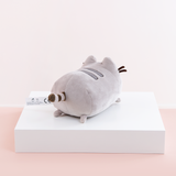 Back quarter view of the Mini Log Squisheen, facing the right, placed on top a square white pedestal in front of a light pink and white background. Pusheen’s behind is just as round as her front. Her striped tail sits directly in the middle of the circle. There are two nub feet at the bottom of the circle, mirroring the ones up front. An information tag sticks out to the left of the tail.