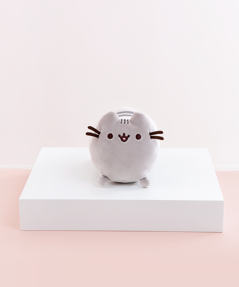 Front view of the Mini Log Squisheen plush, which has been placed on a square white pedestal in front of a light pink and white background. The front of Pusheen is a perfect circle, save for her two ears poking out on top. Pusheen’s face and whiskers are located closer to the top, while her nub feet are at the very bottom. Pusheen’s back stripes are partially visible through the gap between Pusheen’s ears.