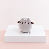 Front view of the Mini Log Squisheen plush, which has been placed on a square white pedestal in front of a light pink and white background. The front of Pusheen is a perfect circle, save for her two ears poking out on top. Pusheen’s face and whiskers are located closer to the top, while her nub feet are at the very bottom. Pusheen’s back stripes are partially visible through the gap between Pusheen’s ears.