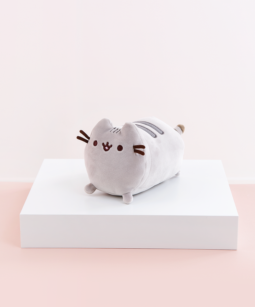 Quarter profile view of the Mini Log Squisheen, facing the left, placed on top a square white pedestal in front of a light pink and white background. Pusheen’s body is shaped like a log, with two dark grey stripes on top of her back, and her back feet and tail partially peeking out from her behind.