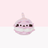 The Macaron Mini Squisheen in front of white and pink background. The plush is Pusheen as a light purple Macaron, laying on her stomach. In the middle of Pusheen is a jagged skirt of light purple fabric and white curved dots of buttercream filling. All of Pusheen’s stripes are pink and on top of her back two stripes. Pusheen’s striped tail sticks out from behind.
