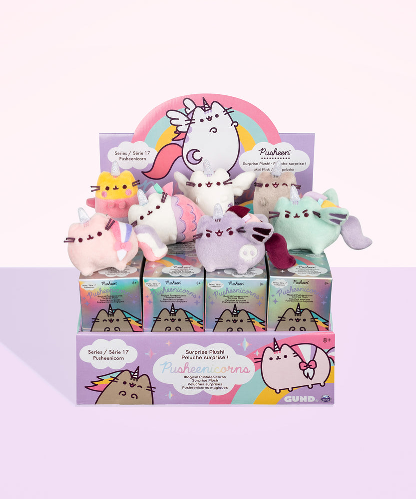All of the Magical Pusheenicorn Surprise Plush resting on top of a cardboard display case holding the surprise box. The individual surprise boxes are iridescent, with an illustration of a classic Pusheenicorn winking. The box display is purple, featuring rainbows, clouds, and illustrations of various Pusheenicorns. The top of the display features Super Pusheenicorn standing proudly underneath a rainbow, her respective surprise plush underneath her.