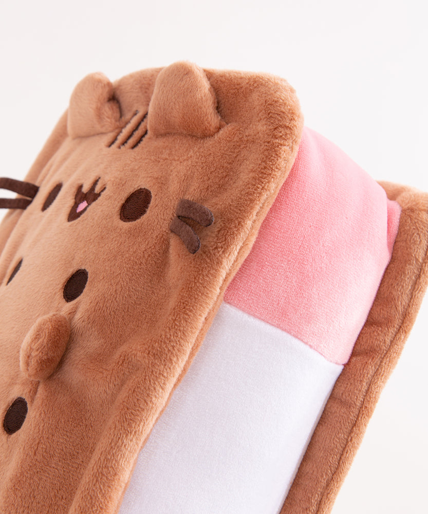 Close-up view of Pusheen Ice Cream Sandwich Squisheen. Pusheen takes the form of an ice cream treat and her classic ears, eyes, stripes, mouth, and whiskers are embroidered at the end of the desert plush. The inside “sandwich” portion is pink, white, and light green. 