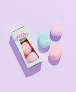 Sponge trio features an embossed Pusheen the Cat on each sponge. Sponges and accompanying packaging lay atop a purple surface. The egg-shaped sponges come to a point at the top of the sponge. The packaging box features a UFO graphic of Pastel Pusheens.