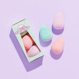 Sponge trio features an embossed Pusheen the Cat on each sponge. Sponges and accompanying packaging lay atop a purple surface. The egg-shaped sponges come to a point at the top of the sponge. The packaging box features a UFO graphic of Pastel Pusheens.