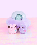 The mini Pink and lilac plush in front of their ship, with the mini mint Pusheen tucked into the window of the ship’s top silver dome. The plush is on top of a holographic floor with a light purple background.