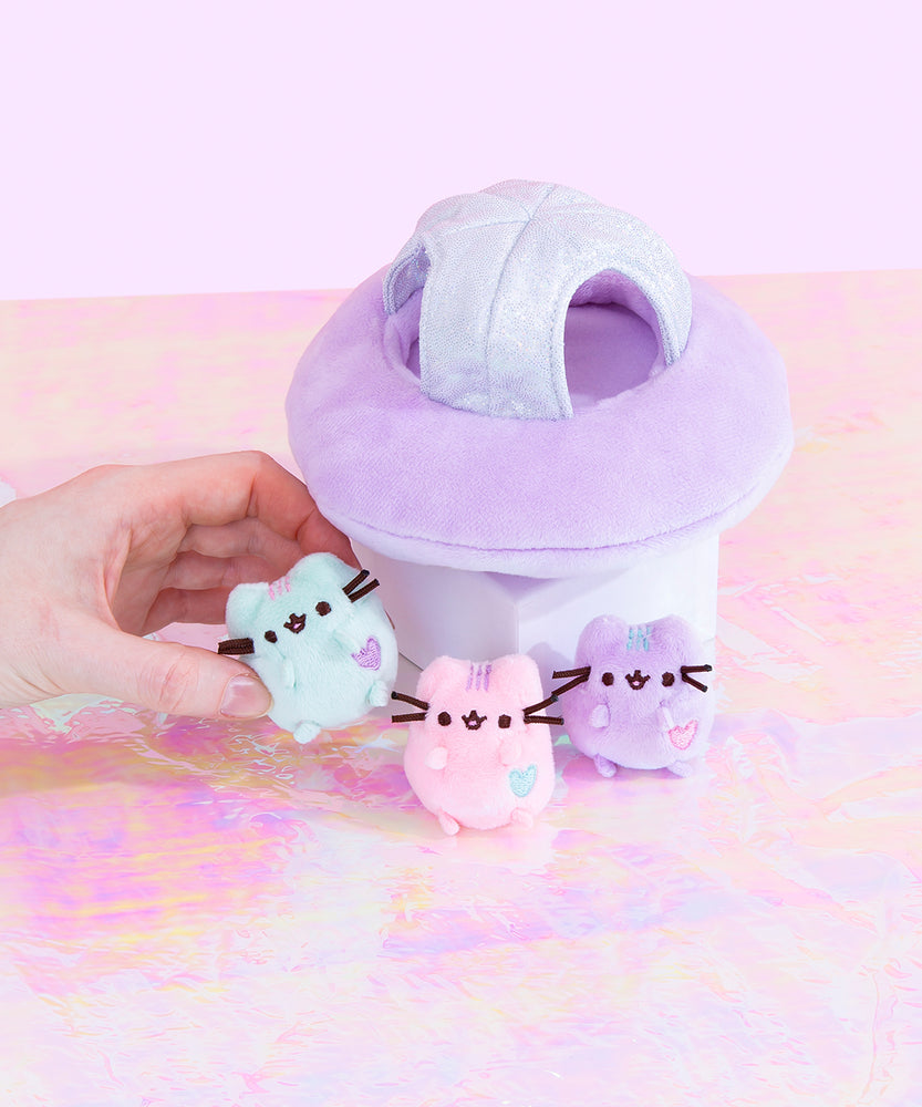 The Mini Pastel Pusheen plush on top of a holographic surface, their ship resting behind them on a pedestal. A model’s hand kidnaps the Mini Mint Pastel Pusheen on the left. The Mint Pusheen has pink stripes and a purple heart, Pink Pusheen has purple stripes and a green heart, and Lilac Pusheen has green stripes and a pink heart.