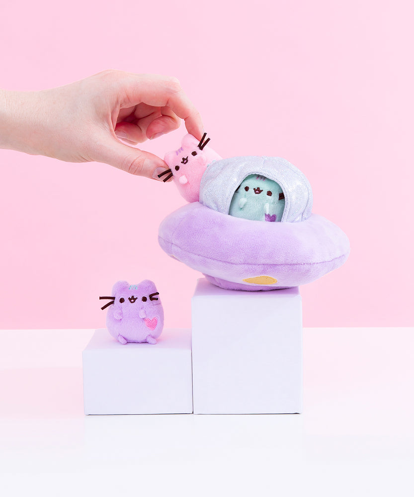 The Pastel Pusheen UFO Collector Set plush arranged on top of white pedestals, a hand taking the mini pastel pink Pusheen out of the UFO. The UFO is a circular purple plush, with a small sparkly silver dome on top with mini windows that the mini pastel mint Pusheen is peeking out of. The mini lilac pastel Pusheen sits on the slightly lower pedestal from the UFO. 