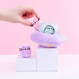 The Pastel Pusheen UFO Collector Set plush arranged on top of white pedestals, a hand taking the mini pastel pink Pusheen out of the UFO. The UFO is a circular purple plush, with a small sparkly silver dome on top with mini windows that the mini pastel mint Pusheen is peeking out of. The mini lilac pastel Pusheen sits on the slightly lower pedestal from the UFO. 