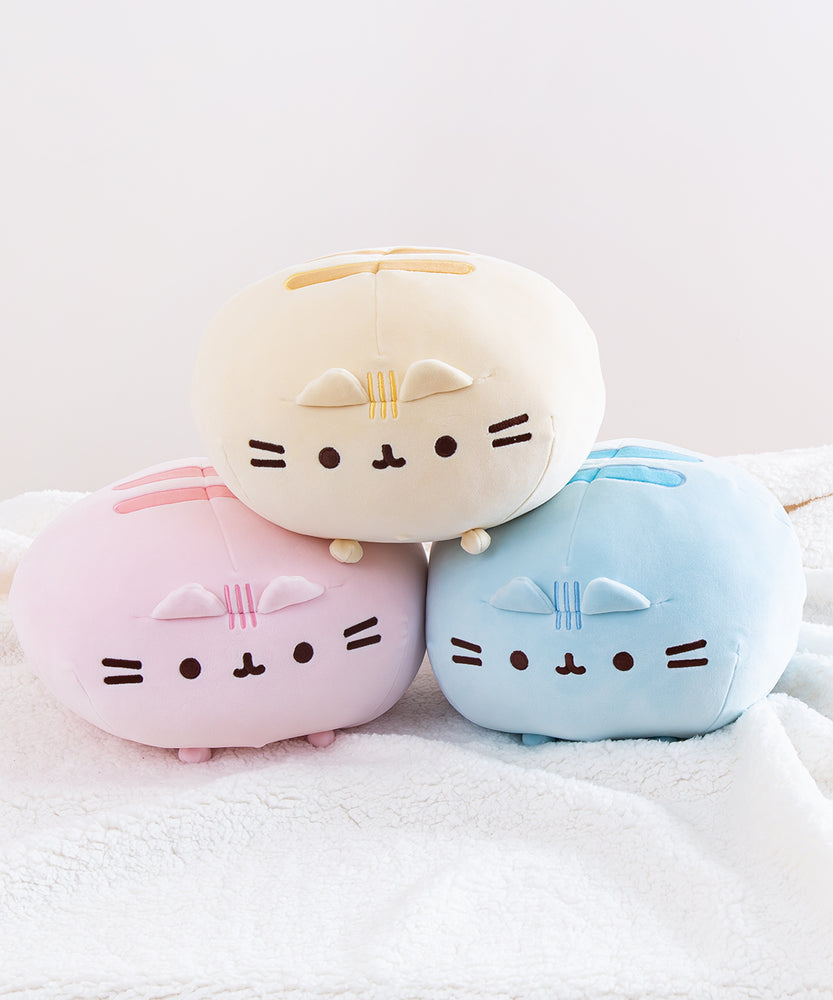 A trio of Round Squisheen plush stacked up in the shape of a triangle, resting on top a fluffy white blanket. All the plush are round, with little stub feet at the bottom, two embroidered stripes on the top, and an embroidered face, whiskers, and head stripes in the middle, with two plush triangle ears sticking out. The pink and blue plush are at the bottom, while the yellow plush sitting on top of them. 