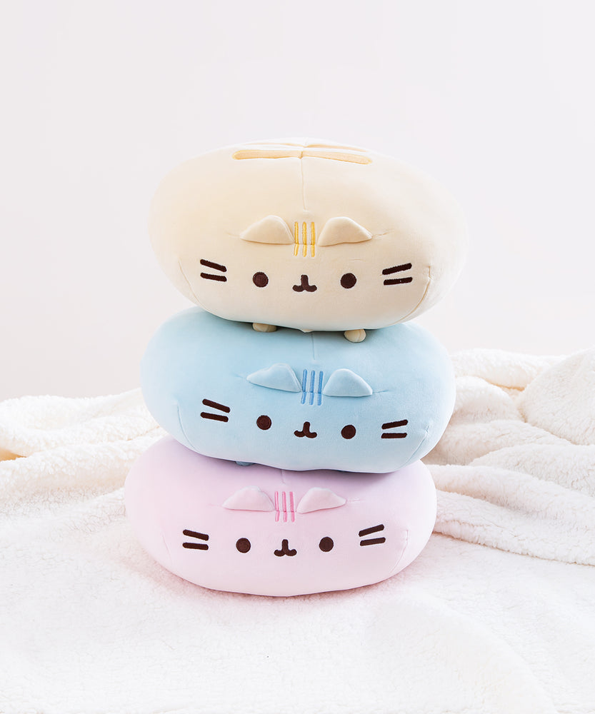 The three Round Squisheen Plush stacked on top of one another, facing the camera and sitting on top a white fluffy blanket. The pink plush is at the bottom, blue in the middle, and yellow on the top. The pink and blue plush are squished down enough to look as if they have no feet, but the yellow round Pusheen still has her feet.