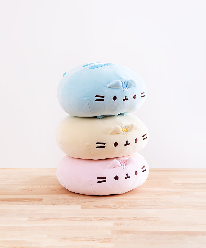 The three Round Squisheen Plush stacked on top of one another, sitting on top a wooden floor in front of a white background. The pink plush is at the bottom, the yellow in the middle, and blue on the top. The pink and yellow plush are squished down enough to look as if they have no feet, but the blue round Pusheen still has her feet.