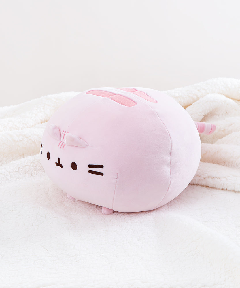The pink Round Squisheen Plush sitting alone in a quarter profile in a sea of a fluffy white blanket.