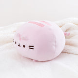 The pink Round Squisheen Plush sitting alone in a quarter profile in a sea of a fluffy white blanket.