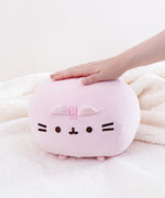 Front view of the Pink Round Squisheen sitting on top a fluffy white blanket, a model’s hand sticking out from the right to rest on top of the plush.