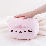 Front view of the Pink Round Squisheen sitting on top a fluffy white blanket, a model’s hand sticking out from the right to rest on top of the plush.