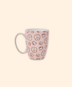 Front view of the Pusheen Pink Donuts & Coffee Mug. The light pink mug has a white handle and interior walls. The outside of the mug features a repeating pattern of Pusheen the Cat as a glazed donut with rainbow sprinkles surrounded by coffee cups and rainbow glazed donuts.