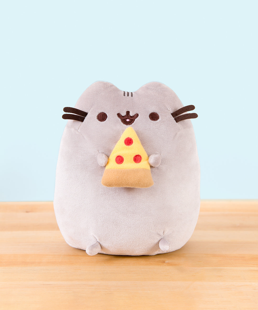 Pusheen sitting upright, mouth open, her two top nub paws holding a plush pepperoni pizza slice, and her bottom nub feet rest at the bottom.