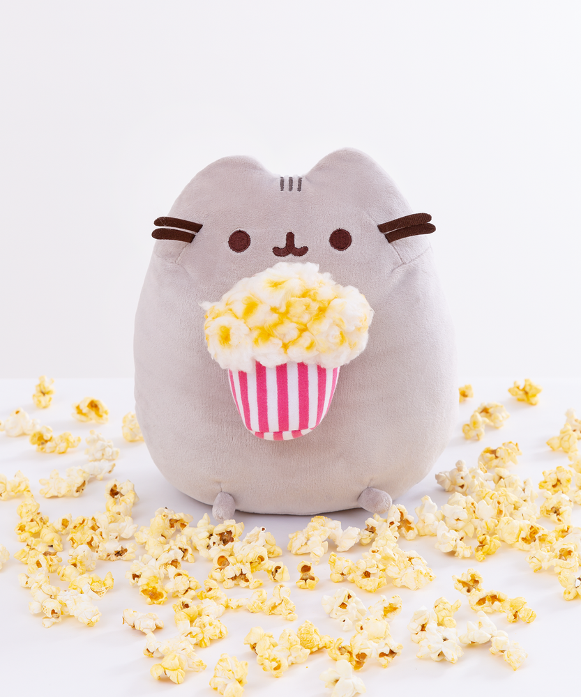 Pusheen sitting upright, her two top nub paws holding a plush striped bag of popcorn, and her bottom nub feet rest at the bottom. The popcorn in the plush basket is made of a different material that resembles cotton balls, and has butter slathered all over it. The plush is surrounded by loose pieces of popcorn.