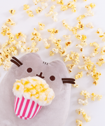 The Popcorn Pusheen Plush peeking out from the lower left corner, loose popcorn pieces strewn all above her. 
