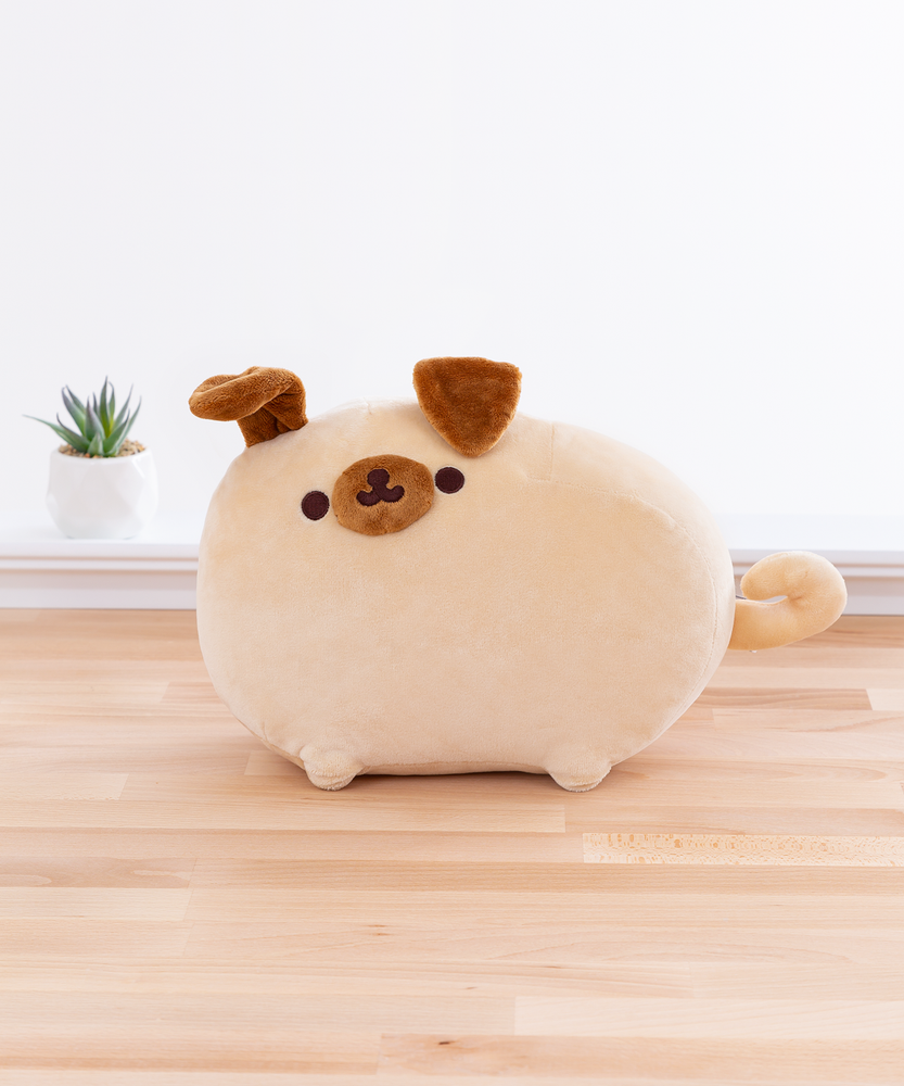 The Pugsheen plush in front of a white wall on top of a wooden floor, and a potted succulent can be seen to the plush’s left. Pugsheen’s left ear is slightly raised, as if something has caught her attention. The inside of her ear is the same brown fabric as the rest of the ear and her snout.