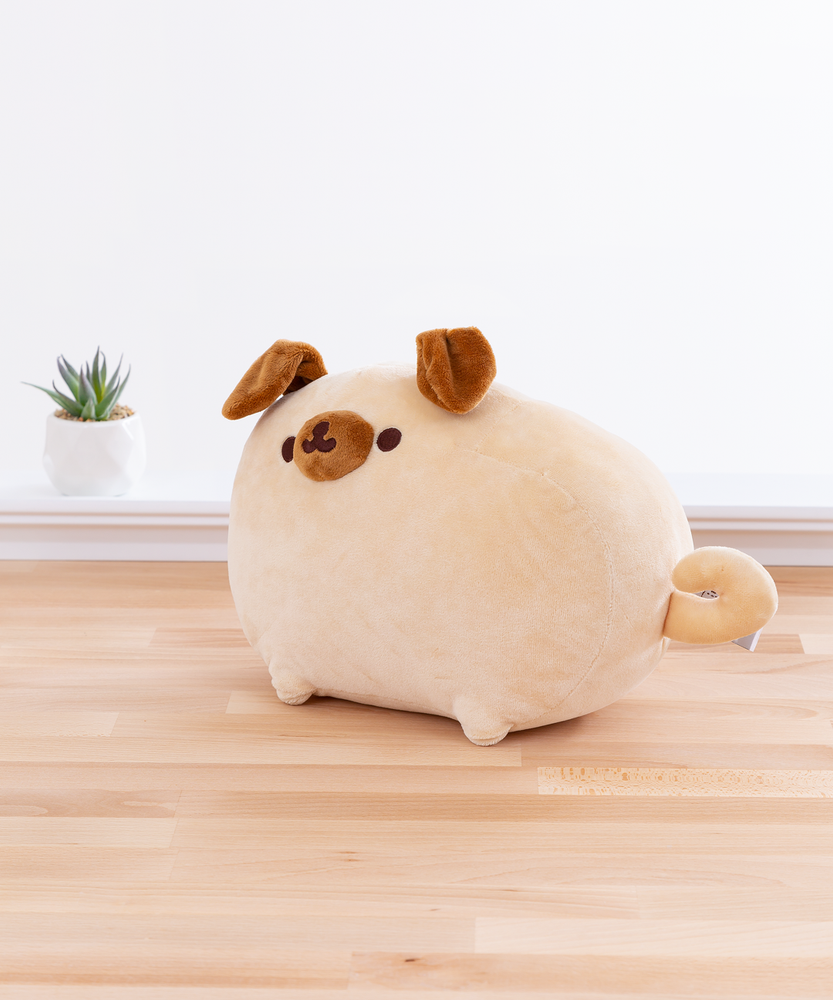 Quarter view of the Pugsheen plush facing the left plush in front of a white wall on top of a wooden floor, with a potted succulent to the plush’s left. Pugsheen’s curly tail covers the plush’s information tag. Pugsheen’s brown snout sticks out from the rest of the plush.