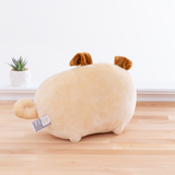 Back view of the Pusheen Plush in front of a white wall on top of a wooden floor, with a potted succulent to the plush’s left. There are no details on the plush’s back, and the information tag on the plush is nearby the curly tail. The back view makes it more obvious Pugsheen’s ears are folded over.
