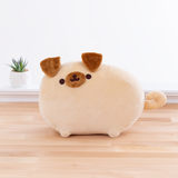 Light brown round dog with a circular brown snout, droopy brown ears that look like triangles, and a curly tail. Pugsheen has the same embroidered dark brown dot eyes and cat mouth as Pusheen the Cat does, and is standing in the same pose as the default Pusheen pose. The plush is in front of a white wall on top of a wooden floor, and a potted succulent can be seen to the left of the plush..