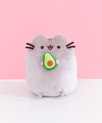 Front view of Pusheen Avocado Plush. Pusheen the Cat is shown with her classic brown eyes, mouth, and whiskers. Pusheen holds a cut-in-half, two-toned green avocado with a light brown pit.