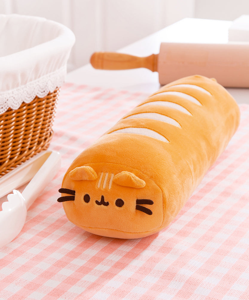 Front view of Pusheen Baguette Squisheen. The elongated brown plush is in the shape of French bread or a baguette. The front of the plush features brown embroidery details with light brown cat ears extending off the top. On the top of the squisheen are cream-colored lines to mimic bake lines. The plush sits on a pink and white surface surrounded by a breadbasket, rolling pin, and cream tongs.  