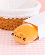 This view shows the baguette plush peeking out of a white bread bag. The golden plush sits on a white and pink checkered surface.  