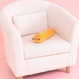 Pusheen Baguette Squisheen sits on a cream chair. The golden baguette-shaped plush has brown embroidery features for Pusheen's eyes, mouth, and whiskers. 