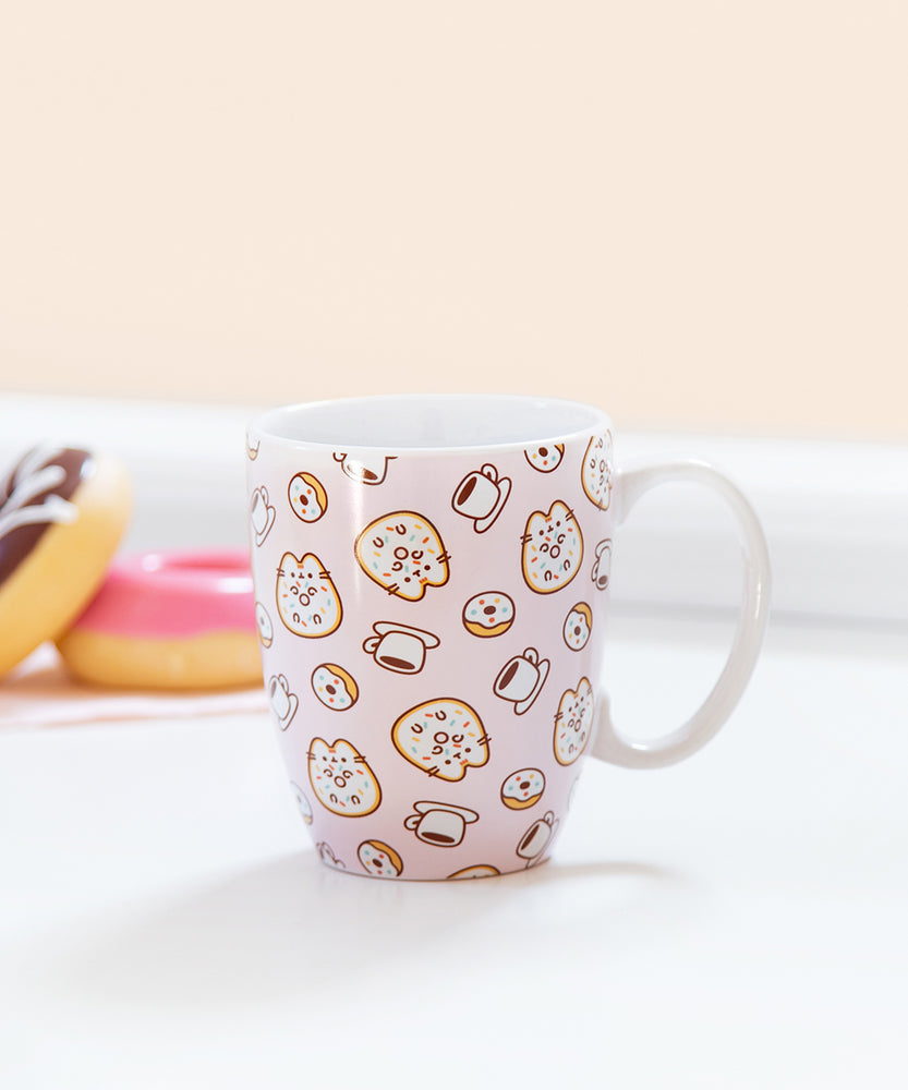 Side view of Pusheen Pink Donuts & Coffee Mug. The white mug has a printed pattern of Pusheen as a glazed donut with coffee and more donuts. The light pink background mug sits on a white surface with donuts faded out in the background.