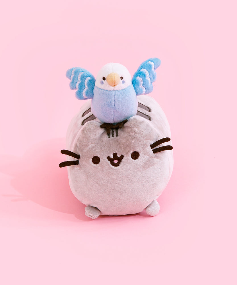 Plush of Bo sitting on Pusheen's head. Bo is a light blue parakeet with a white head, yellow beak, dot eyes and blue dots on her cheek. Bo's darker blue wings are spread out wide with three rows of white feathers, and her dark brown feet are perched in between Pusheens' ears. Pusheen is shaped like a log, laying on her stomach and her paws laying out from the bottom.