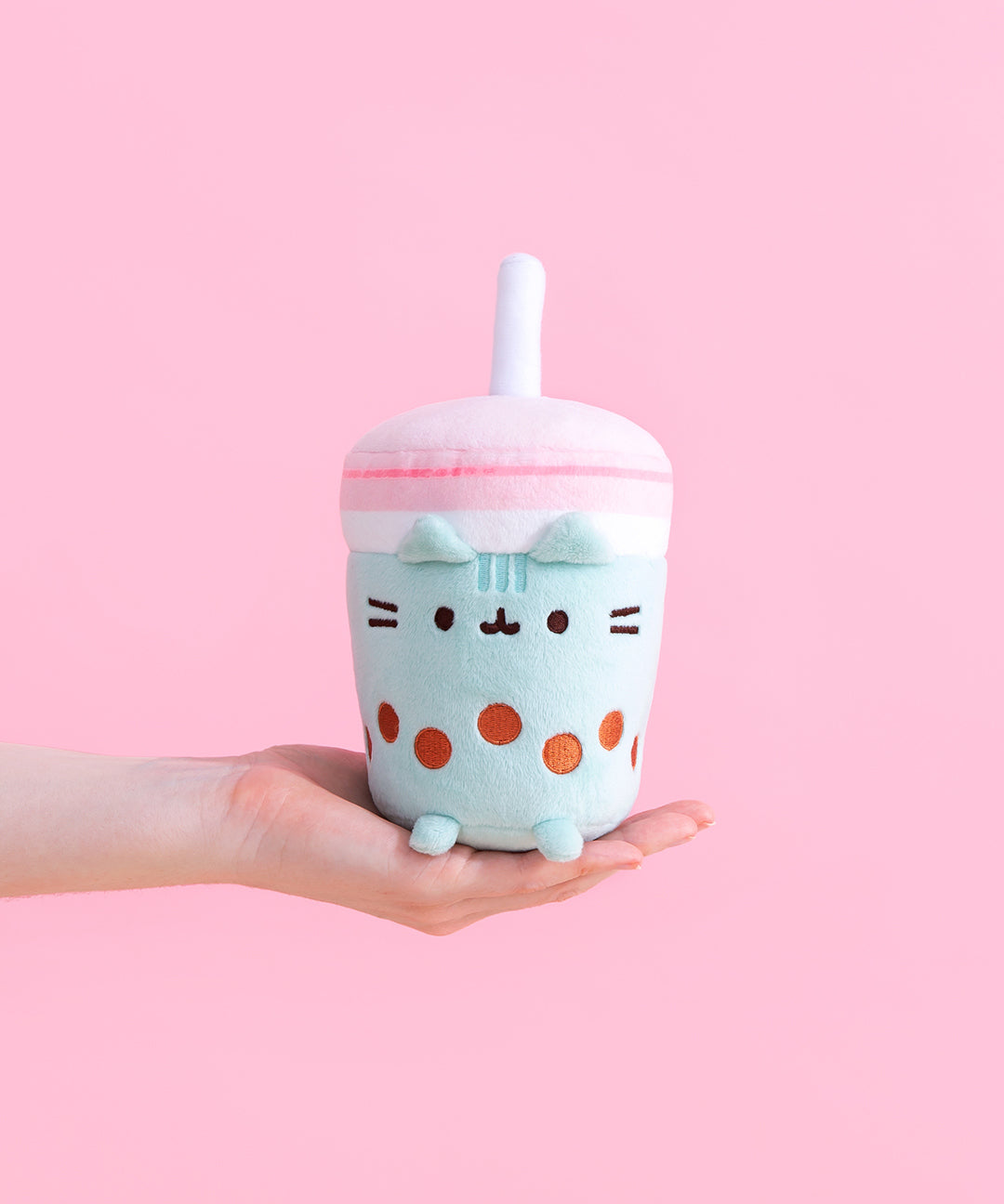 Model’s hand holding out the Pusheen Boba Tea Sips Plush in front of a pink background Pusheen is in the shape of a cylinder cup, with a pink lid and a non-removeable plush straw. The body of the cup is white and mint green, featuring an embroider of Pusheen’s face and whiskers alongside a string of brown boba pearls. Pusheen’s plush ears stick out from the rim of the mint drink, and plush feet stick out up front. 