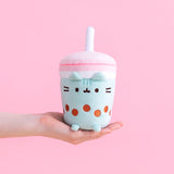 Model’s hand holding out the Pusheen Boba Tea Sips Plush in front of a pink background Pusheen is in the shape of a cylinder cup, with a pink lid and a non-removeable plush straw. The body of the cup is white and mint green, featuring an embroider of Pusheen’s face and whiskers alongside a string of brown boba pearls. Pusheen’s plush ears stick out from the rim of the mint drink, and plush feet stick out up front. 