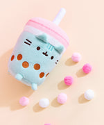 Close-up view of the Boba Tea Sips Plush. On a light-yellow surface, the plush is laid down on its back with its feet up and is surrounded by multi-color pink pom-poms. The bottom of the plush is flat, like an actual cup. 